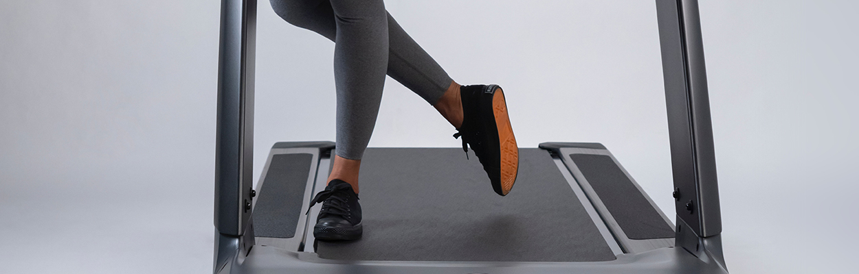 A woman standing on a treadmill, the photo is from her knees down. She is wearing grey leggings and black sneakers with one foot flat and the other leg crossed behind her with the toe pointed.